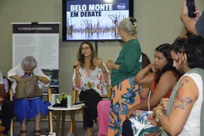Uma mulher sentada no centro, falando ao microfone durante um debate sobre Belo Monte, exibido em um monitor ao fundo. Várias mulheres assistem, com uma senhora em pé à direita.