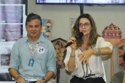 Uma mulher fala ao microfone em um evento, sentada ao lado de um homem. Painéis com textos e desenhos indígenas estão atrás.