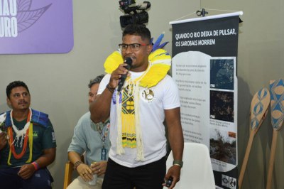 Um homem indígena, com cocar amarelo e branco, fala ao microfone em um evento. Um banner sobre o Rio Xingu está à direita.