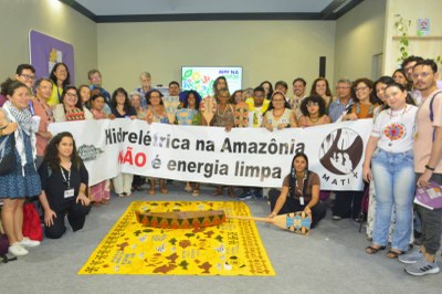 Um grupo de pessoas exibe uma faixa: "Hidrelétrica na Amazônia não é energia limpa". Um tapete e um remo indígena estão em primeiro plano.