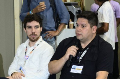 Dois homens em um painel, o da frente com camisa polo preta e crachá, falando ao microfone; o segundo com camisa branca.