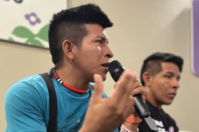 Dois homens sentados, sendo que o primeiro usa camisa azul e fala ao microfone