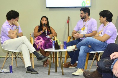 Foto do debate com quatro pessoas sentadas em círculo com uma mesa redonda pequena ao centro; três homens vestem uma camisa lilás e um indígena fala ao microfone 