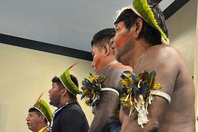 Quatro homens indígenas perfilados com pintura facial, cocares e adereços de penas no braço direito, em ambiente interno.