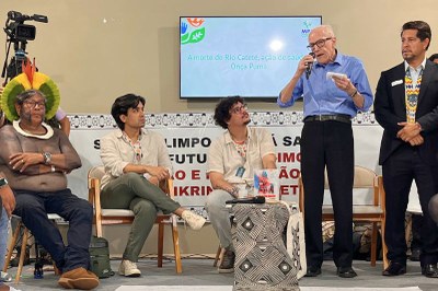 Cinco homens no palco, três sentados e dois em pé, com um projetor exibindo "A morte do Rio Catete, ação de saúde Onça Puma".
