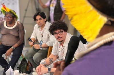 Três homens no centro, um indígena, um jovem com microfone, e um de óculos, sendo observados por alguém em primeiro plano.