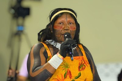 Mulher indígena com pintura corporal, vestido amarelo, colar e luva branca de crochê, falando ao microfone.