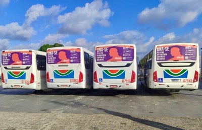 A imagem anexa mostra a parte traseira de quatro ônibus de transporte público estacionados lado a lado, em um pátio aberto sob um céu azul claro com nuvens esparsas. Cada ônibus exibe um "busdoor" idêntico, que é o adesivo promocional afixado na janela traseira. Este adesivo é o mesmo banner da campanha "UNA-SE PELO FIM DA VIOLÊNCIA CONTRA AS MULHERES", com fundo roxo e a silhueta laranja de uma mulher, juntamente com os logotipos das entidades apoiadoras como o MPF, MPT e a SINTUR. A repetição do anúncio nos quatro veículos, que são brancos com detalhes coloridos e números de frota visíveis (como 07023, 07060, 07117 e 07043), sugere que a campanha está sendo veiculada em larga escala para conscientização pública na cidade.