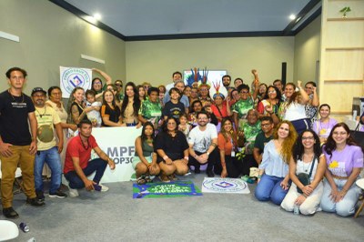 Grande grupo de pessoas, incluindo adultos e crianças, posando em frente a um banner, algumas com cartazes no chão.