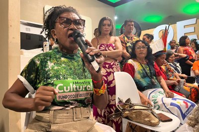 Mulher sentada com camisa estampada verde e branca, falando em um microfone, com outras pessoas em segundo plano.