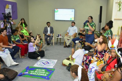 Pessoas sentadas em círculo em evento, algumas em cadeiras e outras no chão, duas pessoas em destaque no centro em cadeiras, e uma mulher em pé falando ao microfone, com um telão ao fundo.