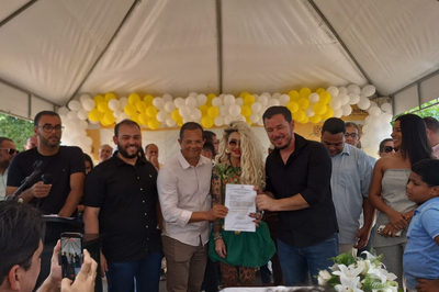 Cerimônia pública ao ar livre, com quatro pessoas, incluindo uma mulher loira e tatuada, assinando um documento sob uma tenda decorada com balões amarelos e brancos.