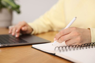 Imagem em detalhe de uma pessoa com casaco amarelo escrevendo em um caderno com uma mão, enquanto a outra está no notebook ao lado