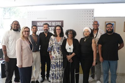 Foto dos artistas em frente a um backdrop com o nome da exposição junto com as procuradoras Ana Chiodelli e Daniele Escobar