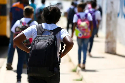 Foto: Marcelo Camargo/Agência Brasil Jovem de costas, usando camiseta branca e mochila escura, em primeiro plano, caminhando em direção a outros estudantes, também com mochilas. Eles estão em uma rua ou calçada sob luz intensa.