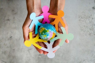 Duas mãos seguram recortes de papel coloridos em forma de pessoas em círculo ao redor de um pequeno globo terrestre