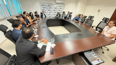 A terceira imagem mostra uma visão ampla da mesma sala de reuniões, com diversos participantes posicionados em ambos os lados da mesa. No centro, estão os representantes que conduzem o diálogo, enquanto os demais acompanham com atenção, alguns tomando notas. O cenário institucional e organizado reforça o caráter técnico e colaborativo do encontro.