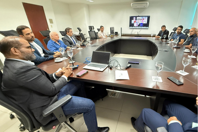 A foto mostra uma reunião na sala de conferências da Procuradoria da República em Alagoas (MPF/AL). Os participantes estão dispostos ao redor de uma mesa em formato de “U”, com notebooks, celulares, copos e xícaras de café à frente. No fundo da sala, um monitor exibe pessoas participando de forma virtual. O ambiente é formal e bem iluminado, com um clima de atenção e diálogo institucional.