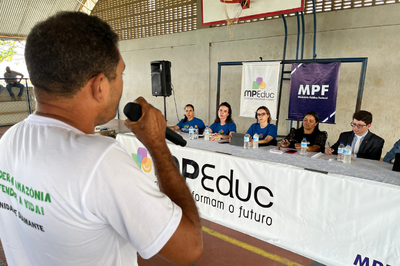 Um homem, de costas, em primeiro plano, fala ao microfone em um evento. Seis pessoas estão sentadas à mesa, olhando pra ele. Atrás delas há dois banners com as logos do MPF e do MPeduc.