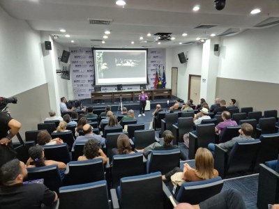 Momento da audiência pública. Imagem: Comunicação MPF A imagem mostra um auditório com várias pessoas sentadas em cadeiras acolchoadas de cor azul, todas voltadas para um palco onde ocorre uma apresentação. No palco, há um palestrante em pé diante de uma tela grande que exibe uma projeção. Atrás dele, vê-se um painel com a sigla MPF repetida diversas vezes, indicando que o evento está relacionado ao Ministério Público Federal.