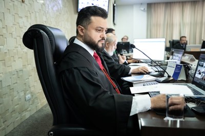 Fotos: Secom TRE e Ascom MPF/GO A imagem mostra um homem vestido com toga e gravata vermelha sentado em uma cadeira de couro preta, em uma mesa de tribunal ou plenário. Ele está concentrado em frente a um notebook, com documentos à sua frente e um microfone posicionado para fala. Ao fundo, outros profissionais também trajando togas trabalham em computadores, sugerindo que a sessão é híbrida, com participantes presenciais e virtuais — evidenciado pelas telas exibindo videoconferências.