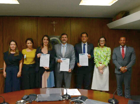 Fotografia colorida em ambiente corporativo, possivelmente uma sala de reuniões ou gabinete com painéis de madeira marrom escuro ao fundo. Sete pessoas (quatro mulheres e três homens) estão lado a lado, em pé, atrás de uma grande mesa, todas vestidas em trajes sociais ou executivos. As três pessoas ao centro seguram e exibem papéis (aparentemente, um acordo ou termo) que acabaram de assinar. 