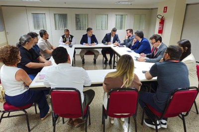 Foto em ambiente interno de uma reunião de trabalho. Homens e mulheres estão sentadas ao redor de uma mesa retangular branca. As pessoas vestem trajes sociais e casuais, e algumas estão usando celulares ou interagindo. No fundo, há janelas horizontais com venezianas e um extintor de incêndio vermelho na parede.