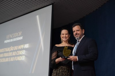 A imagem é uma fotografia do procurador Juraci Guimarães recebendo o troféu do Prêmio Sociedade. O procurador está de terno ao lado de uma mulher de cabelo preso e roupa preta. O prêmio é dourado e se assemelha a um rosto de uma mulher com asas.