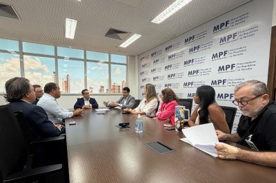Foto: Comunicação/MPF Foto de uma mesa com nove pessoas sentadas ao redor em uma reunião, ao fundo janela com vista da cidade e na parede ao lado, banner com a marca do MPF