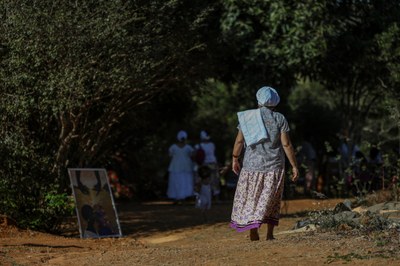 Foto: Marcelo Camargo/Agência Brasil. Foto mostra uma mulher idosa com lenço branco na cabeça caminhando em estrada de terra em direção a um grupo de pessoas de roupas brancas, em uma comunidade tradicional ou quilombo.