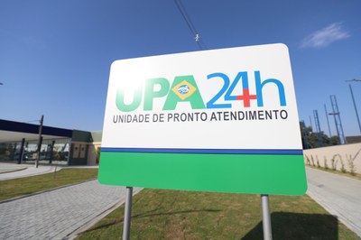 Imagem ilustrativa - Prefeitura de Paranaguá/PR Placa de identificação da UPA 24h (Unidade de Pronto Atendimento), em fundo branco, com a sigla em verde e o número 24h em azul e vermelho. A placa está em um ambiente externo, sob um céu azul claro, com grama verde e um caminho pavimentado. No fundo, à esquerda, aparece o prédio da unidade de saúde.