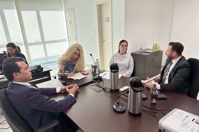Reunião entre o MPF, representado pela procuradora da República Gisele Bleggi (centro-esquerda), e representantes da Iguá, em 18/11/2025, na sede do órgão, em Aracaju (SE). Foto: Comunicação/MPF. A imagem anexa mostra uma reunião de negócios ou profissional em uma sala de conferências bem iluminada, possivelmente um escritório moderno com grandes janelas à esquerda. Em torno de uma mesa de madeira escura e retangular, estão sentadas cinco pessoas. No primeiro plano, à esquerda, um homem de terno azul escuro está sentado, com as mãos cruzadas sobre a mesa, olhando para o lado oposto. À sua frente, um microfone de conferência e duas garrafas térmicas grandes (tipo 'pump pot') se destacam. No centro da mesa, uma pessoa com cabelos loiros cacheados e uma maquiagem marcante, usando uma blusa estampada, está inclinada sobre papéis e um laptop. No lado direito da mesa, duas pessoas estão engajadas na conversa: uma mulher de óculos e blusa cinza claro está sentada ao lado de um homem de terno preto e gravata, que gesticula enquanto fala, sugerindo que ele é o orador naquele momento. Ao fundo, uma quinta pessoa está sentada em uma estação de trabalho separada, olhando para um monitor.