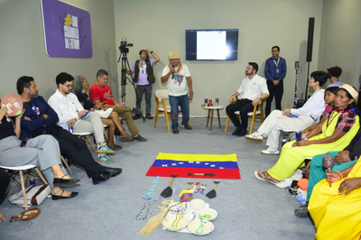 Imagem do estande do MPF no momento dos debates. Um grupo de pessoas sentadas em semicírculo observa um homem em pé no centro, vestindo um cocar. No chão, em frente a ele, está uma bandeira da Venezuela e artefatos indígenas.