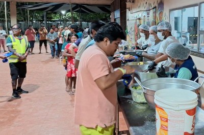 Quatro funcionários para servir o jantar para cerca de 3 mil indígenas na Aldeia COP, na UFPA, em Belém (PA), em 15/11/2025. Foto: MPF. Foto de pessoas em uma fila em um local coberto recebendo comida servida por voluntários em uma bancada com grandes panelas.