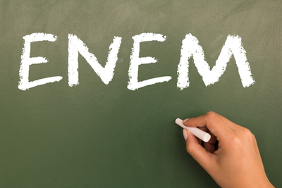 Imagem: Canva.com Imagem de um quadro verde, com uma mão segurando um giz e escrito no quadro a palavra ENEM