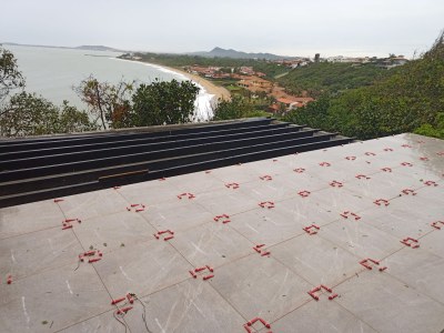 Foto em plano aberto das obras do mirante. A imagem mostra um piso sendo instalado em um terraço com vista para o mar. Em primeiro plano, o piso de cor cinza claro com veios está assentado sobre o contrapiso. Peças plásticas vermelhas, usadas como espaçadores e niveladores, estão distribuídas sobre as juntas das placas em formação de grade. À esquerda, uma estrutura preta em faixas horizontais sugere uma área de deck em construção.

Ao fundo, a paisagem é dominada pela costa: uma faixa de areia clara, a água do mar cinzenta e o horizonte se fundindo sob um céu nublado. Casas de telhados de terracota estão dispostas na colina que margeia a praia, cobertas por vegetação densa.
