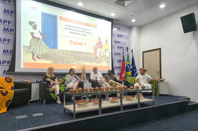 Foto de painel de debate sobre "Aquilombando-se" e diversidade racial do MPF com cinco participantes, incluindo um homem indígena, sentados em frente a uma tela de projeção e uma mesa com cerâmicas artesanais.