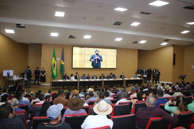 Foto mostra auditório da Assembleia Legislativa com pessoas sentadas, de costas, algumas com chapéus de boiadeiro. Ao fundo, uma mesa com autoridades e no telão a imagem do orador naquele momento, o procurador Gabriel de Amorim.