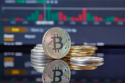 Imagem mostra ilustração que faz referência a uma moeda com o símbolo de bitcoin. Ao fundo, há gráficos desfocados, em referência a movimentações do mercado financeiro