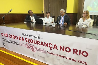 O procurador da República Eduardo Benones (centro) participa do seminário “O Nó da Segurança no Rio", promovido pela ABI, no Rio de Janeiro, em 18/11/2025. Foto: Comunicação/MPF. A imagem registra um momento de um seminário realizado na ABI (Associação Brasileira de Imprensa). Quatro participantes estão sentados à mesa, em frente a uma plateia invisível: três homens e uma mulher. No centro da cena, há uma grande faixa branca que serve como principal elemento visual, exibindo a manchete "O NÓ CEGO DA SEGURANÇA NO RIO" em letras garrafais vermelhas, acompanhada do subtítulo "ESTRATÉGIAS PARA SUPERAR CONFRONTOS E SALVAR VIDAS". A faixa também indica a data do evento: "18 de novembro de 2025". A composição sugere um debate sério e focado em questões cruciais de segurança pública no Rio de Janeiro.