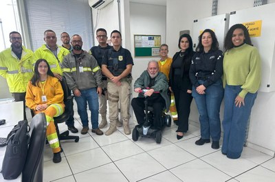 Foto de comunicação do MPF sobre a fiscalização de um posto de pesagem na BR-101 em Araranguá, SC, que mostra um grupo diverso de aproximadamente doze pessoas reunidas em um ambiente interno, que se assemelha a um escritório ou sala de controle. O grupo é composto por homens e mulheres, com alguns vestindo coletes de segurança nas cores amarelo neon e cinza (provavelmente representando a concessionária da rodovia, como a CCR, e/ou equipes técnicas), um homem ao centro em uniforme da Polícia Rodoviária Federal (PRF), um homem mais velho sentado em uma cadeira de rodas motorizada, e algumas mulheres em trajes sociais ou casuais, à direita. A diversidade de vestimentas sugere a participação de diferentes órgãos e entidades na ação de fiscalização, como o Ministério Público Federal (MPF), agentes de segurança e possivelmente a equipe da concessionária da rodovia.