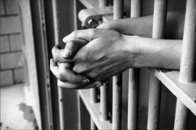 Foto ilustrativa: Canva Imagem em preto e branco de uma cela mostrando em detalhe o braço de um preso com as mãos cruzadas para fora
