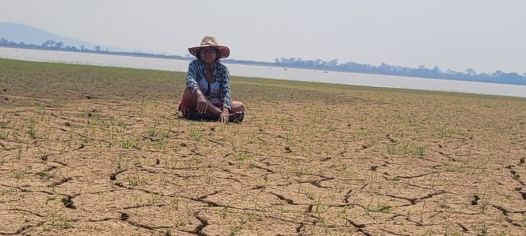 A imagem mostra uma mulher sentada, usando um chapéu e roupas leves, em primeiro plano, sobre um solo completamente rachado e seco, indicando um período de forte seca.