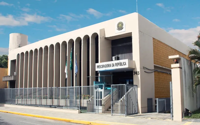 Imagem da fachada do prédio da PR/RN, bege com grades na frente, tendo acima o céu azul e observando-se parte da calçada em frente ao local
