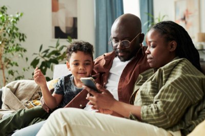 Foto ilustrativa: Pressmaster, via Canva foto de uma família com pai, mãe e filho pequeno, sentada no sofá da sala, olhando para um smartphone.