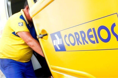 Imagem ilustrativa. Foto: Correios/Divulgação. Fotografia de um trabalhador pegando alguma coisa no interior de uma Kombi amarela, com o nome Correios grafado em letras azuis na lateral do veículo.