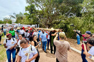 Foto: Arquivo pessoal Cortejo com dezenas de pessoas, algumas carregando uma grande cruz de madeira e outras segurando pequenas cruzes brancas, em ato. Ao fundo, faixa com fotos e árvores.