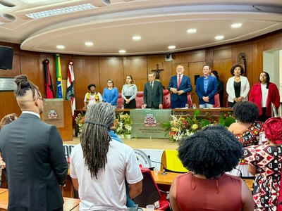 Imagem: Comunicação MPF A imagem mostra uma cerimônia formal no plenário da Câmara Municipal de João Pessoa. Ao fundo, dez pessoas estão em pé atrás de uma longa bancada decorada com arranjos florais.