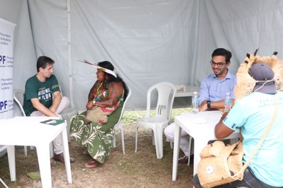 Em uma tenda branca com paredes de lona, duas mesas de plástico branco estão dispostas, cada uma com duas pessoas conversando. À esquerda, o procurador Ramiro Rockenbach conversa com uma mulher indígena. À direita, o procurador Marcos André conversa sorridente com um homem indígena.