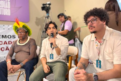 Três homens em primeiro plano, um indígena à esquerda, um homem com microfone no centro e outro com barba e óculos à direita, sentados em um evento.